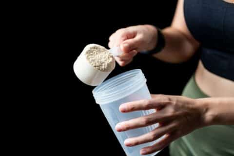 Pessoa segurando um scoop de whey protein em pó e adicionando o suplemento em uma coqueteleira, ilustrando o consumo de proteína na rotina de treino.