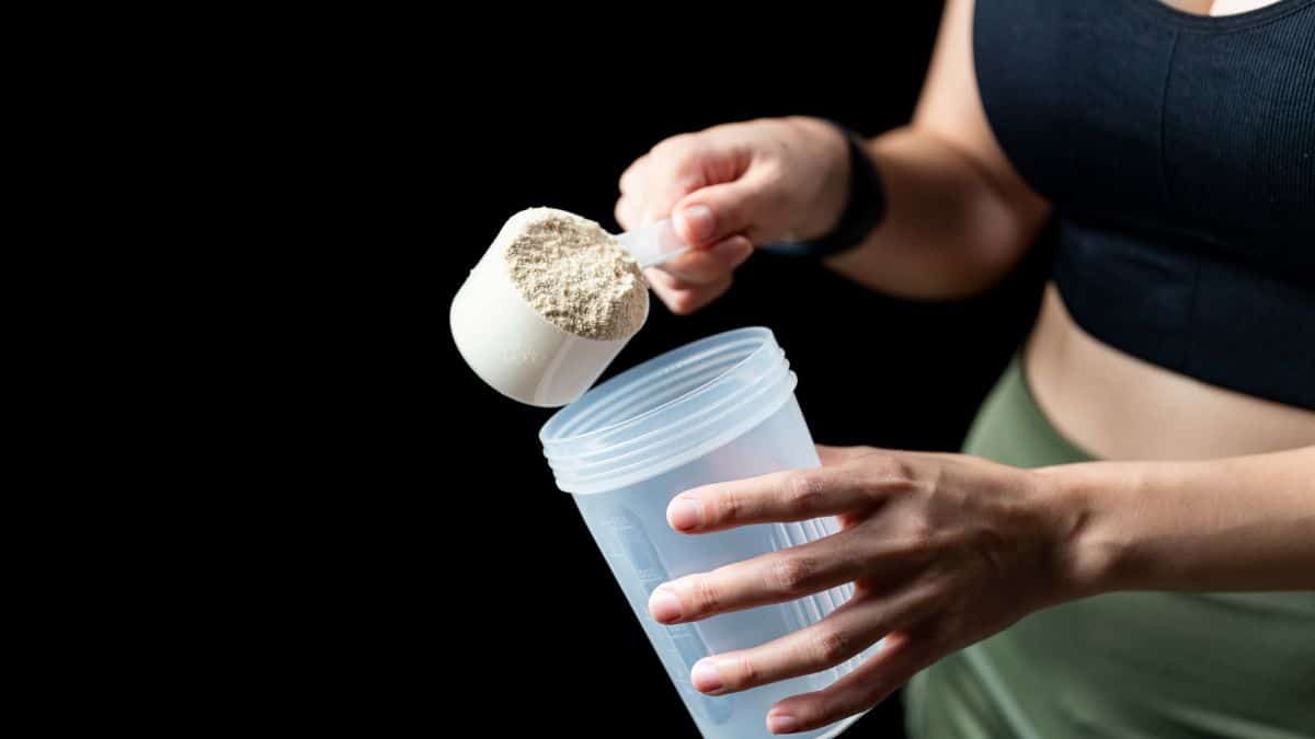 Pessoa segurando um scoop de whey protein em pó e adicionando o suplemento em uma coqueteleira, ilustrando o consumo de proteína na rotina de treino.